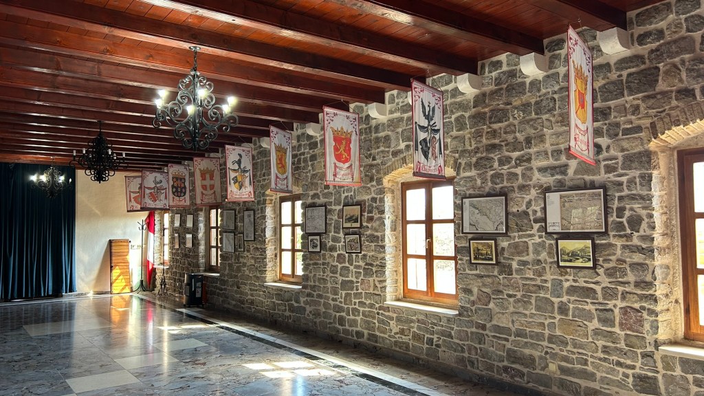 An interior view of a stone-walled hall featuring wooden beams, with banners hanging from the ceiling and framed pictures on the walls. Large windows provide natural light.