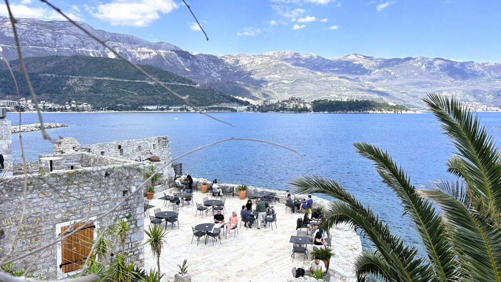 Scenic view of a coastal area with guests seated at outdoor tables, overlooking a tranquil blue sea and mountains in the background.
