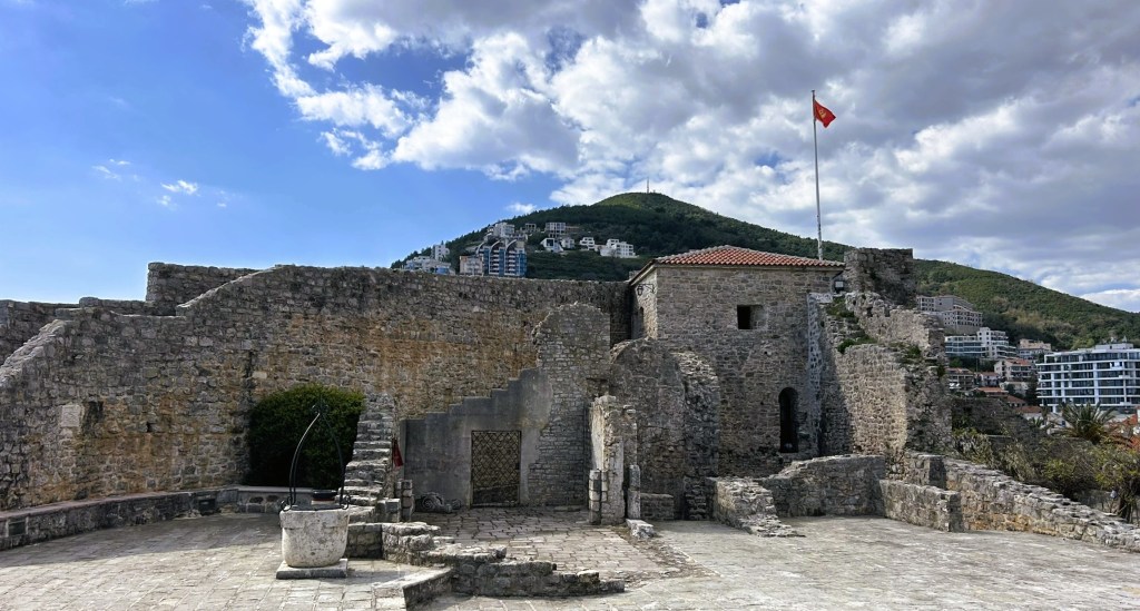 Ruins of a stone fortress with a Turkish flag fluttering, overlooking a green hill and modern buildings in the background under a partly cloudy sky.