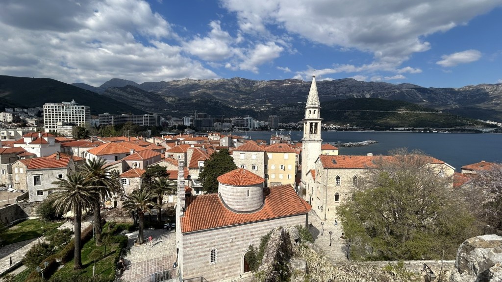 A panoramic view of a coastal town with red-tiled roofs, a steeple, and palm trees, set against mountains and a blue sky with clouds.