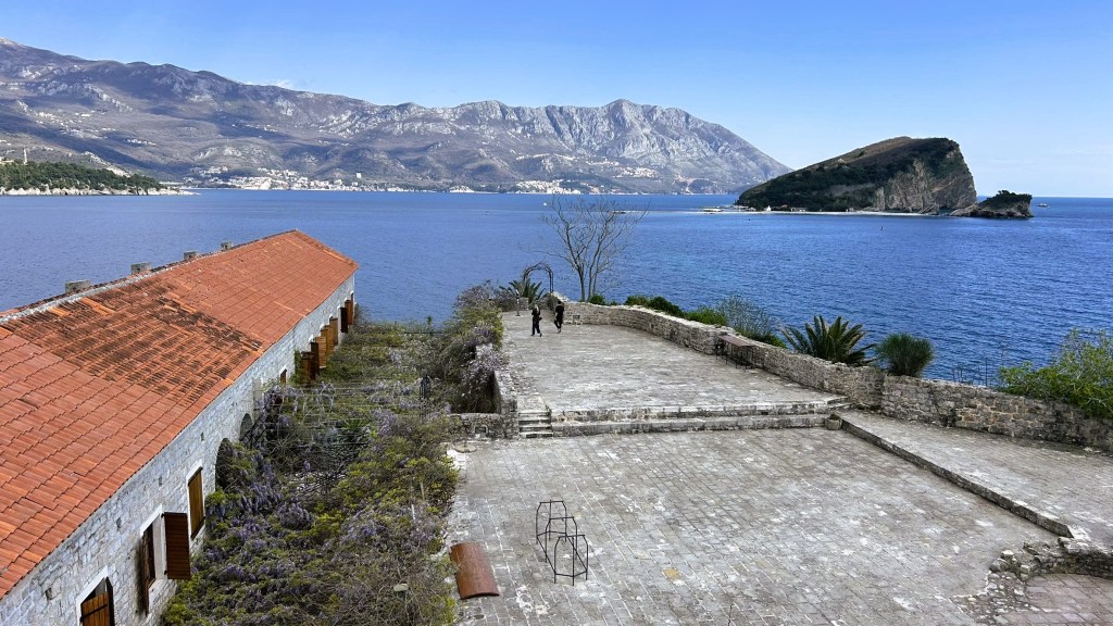 View of a coastal landscape featuring a historical stone building with a red roof, an open plaza, and a rocky island in the distance, surrounded by water and mountains.
