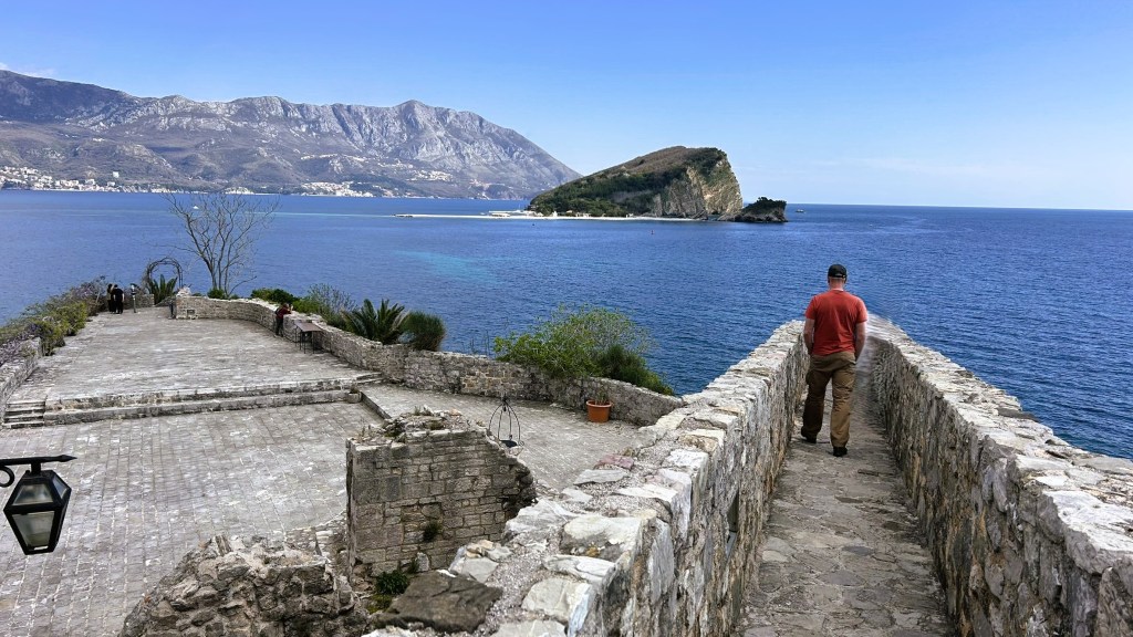 A person walking along a stone pathway with a view of the sea and a small island in the background, surrounded by mountains.