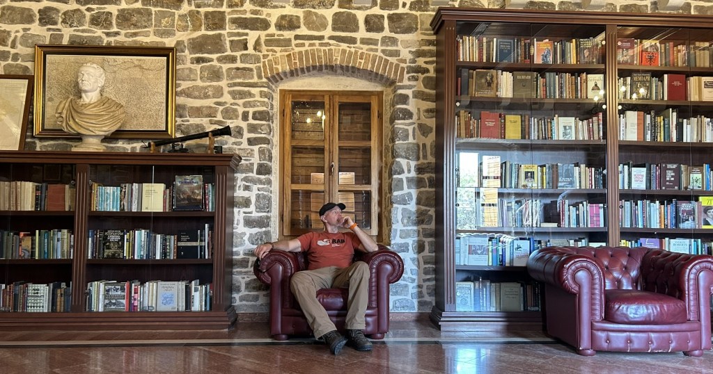 A man sitting in a leather armchair in a library with stone walls, surrounded by bookshelves filled with books, and a bust of a historical figure on the wall.