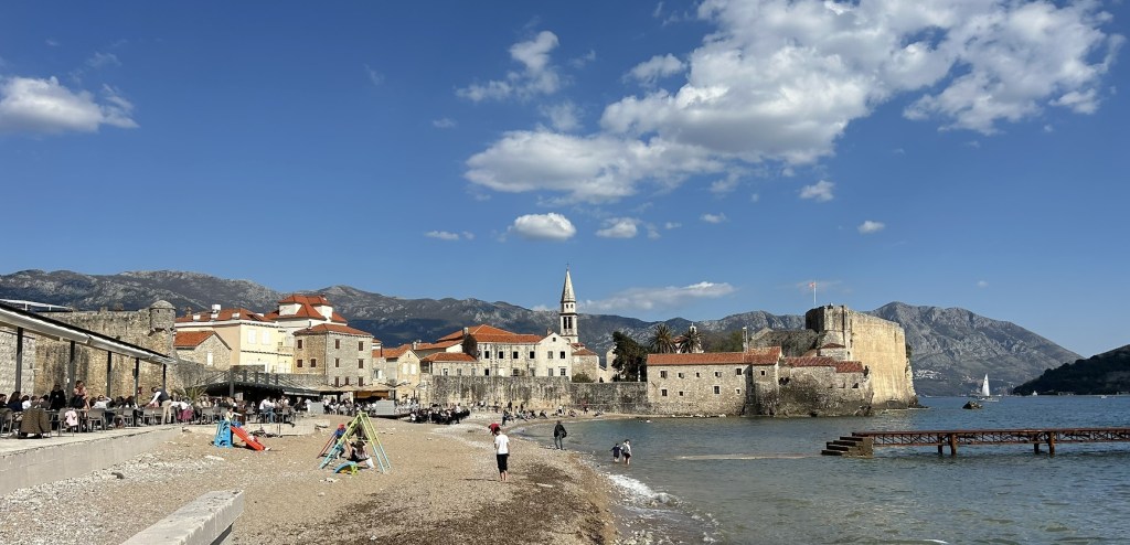 A picturesque beach scene with people enjoying the sun, featuring historic stone buildings with a church steeple in the background, mountains visible under a blue sky with scattered clouds.