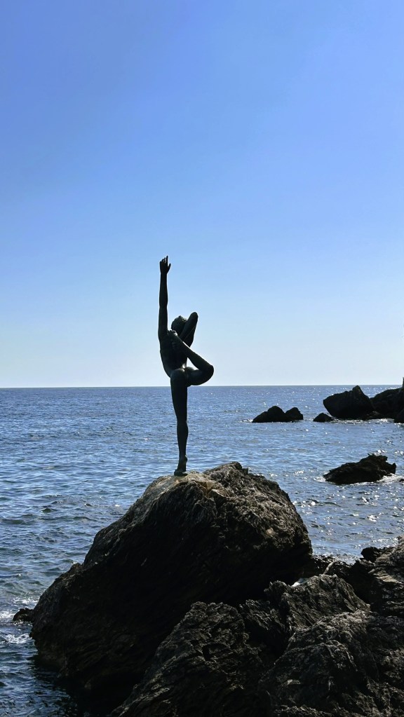 Silhouette of a dancer striking a pose on a rocky outcrop by the sea, with a clear blue sky in the background.