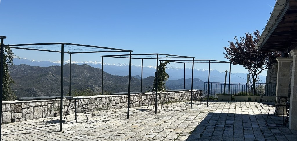 Outdoor terrace with metal frames and chairs, overlooking mountains and clear blue sky.