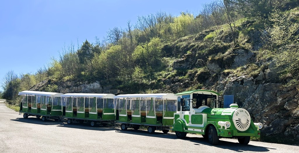 A green tourist train parked on a road, consisting of multiple carriages, set against a backdrop of rocky hills and greenery under a clear blue sky.
