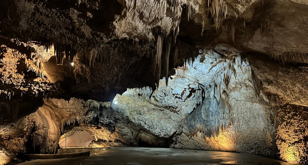 Interior view of a large cave with stalactites and stalagmites, illuminated by soft lighting
