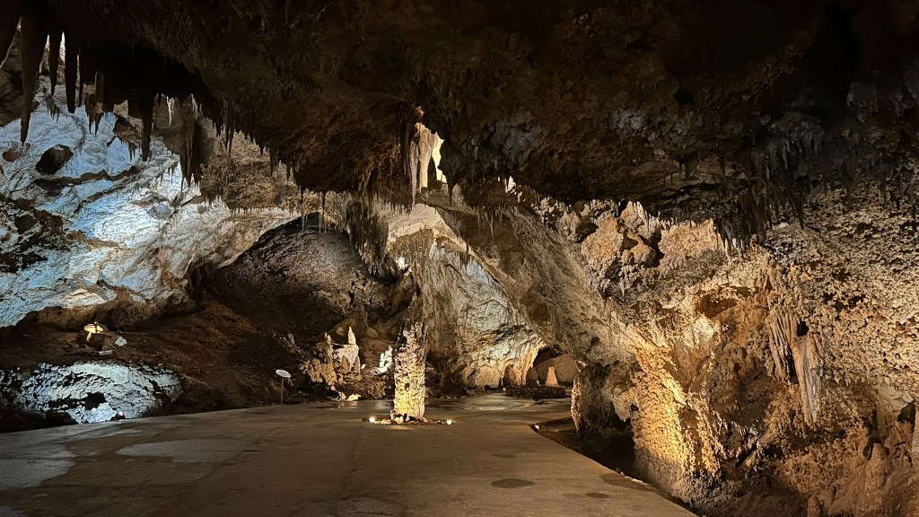 A dark underground cave with stalactites hanging from the ceiling, illuminated by soft lighting, revealing textured rock formations and a clear pathway.