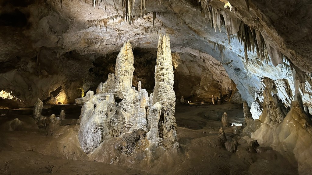 An interior view of a cave featuring tall stalagmites and stalactites, illuminated by soft lighting, showcasing the geological formations and textures of the cave walls.
