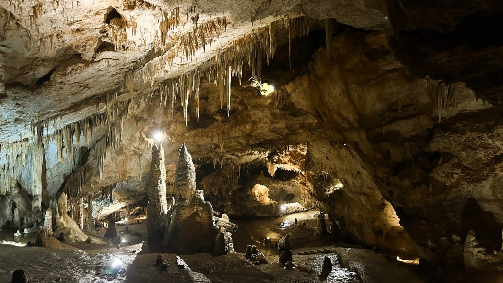 Interior of a large cave featuring stalactites hanging from the ceiling and stalagmites on the ground, with artificial lighting illuminating the rock formations.
