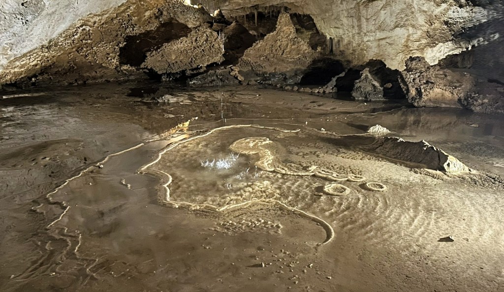 An underground cave with a wet, reflective surface and various rock formations, including stalactites and stalagmites.