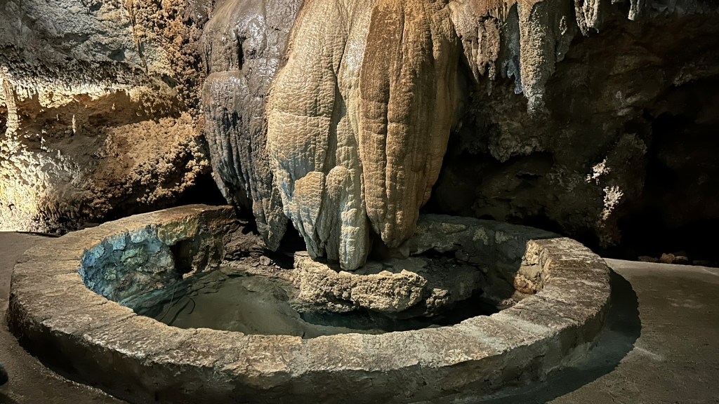 A stone basin with a clear water pool surrounded by a circular stone edge, set within a cave featuring large stalactites and textured rock formations.