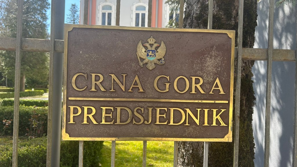 A bronze plaque displaying the words 'CRNA GORA' and 'PREDSJEDNIK', indicating the President of Montenegro, set against a background of greenery and a building.