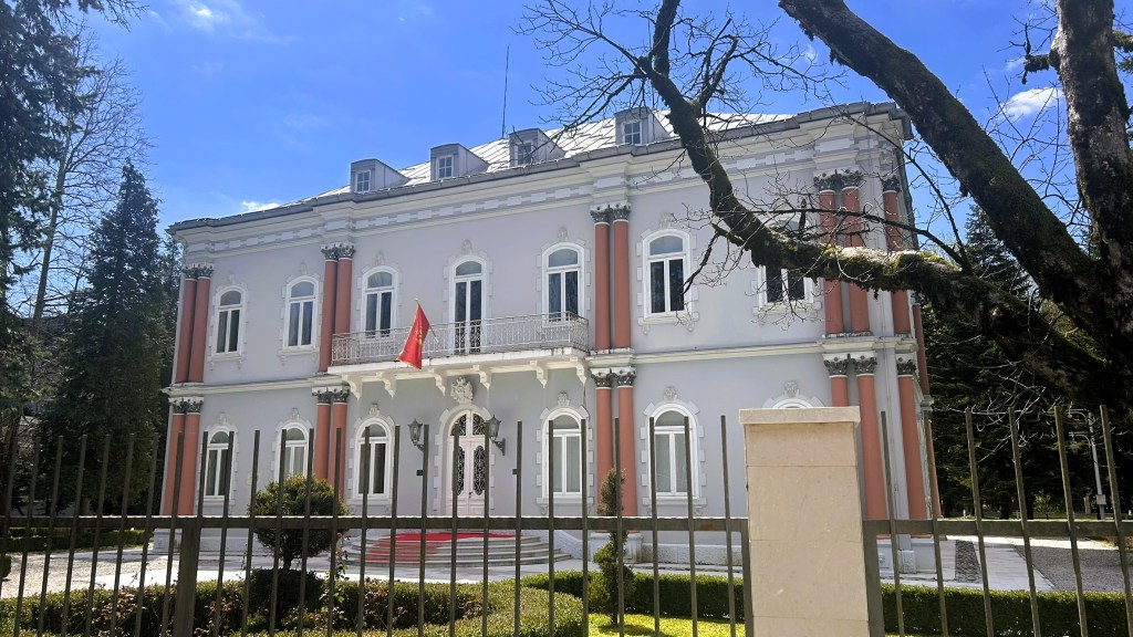A grand, neoclassical building with ornate architecture, featuring pink columns and large windows, surrounded by greenery and a fence, under a clear blue sky.