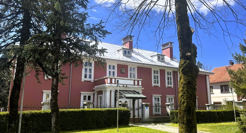 A pink two-storey house with a flat roof and white accents, surrounded by tall trees and green hedges, set against a clear blue sky.