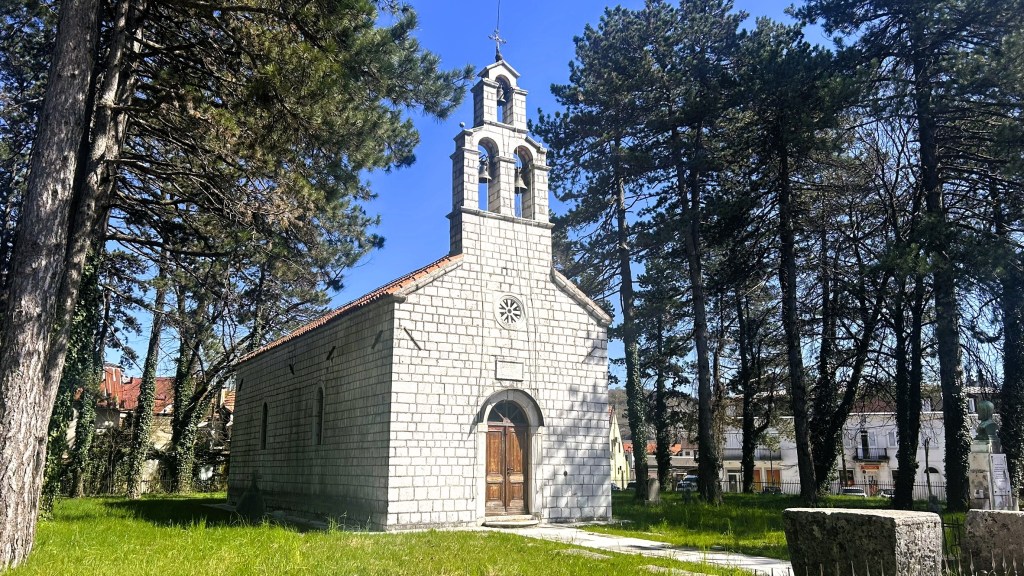 A small stone church surrounded by tall trees and green grass under a clear blue sky.