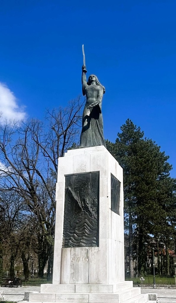 A bronze statue of a woman holding a sword aloft, mounted on a tall stone pedestal, set against a clear blue sky with trees in the background.