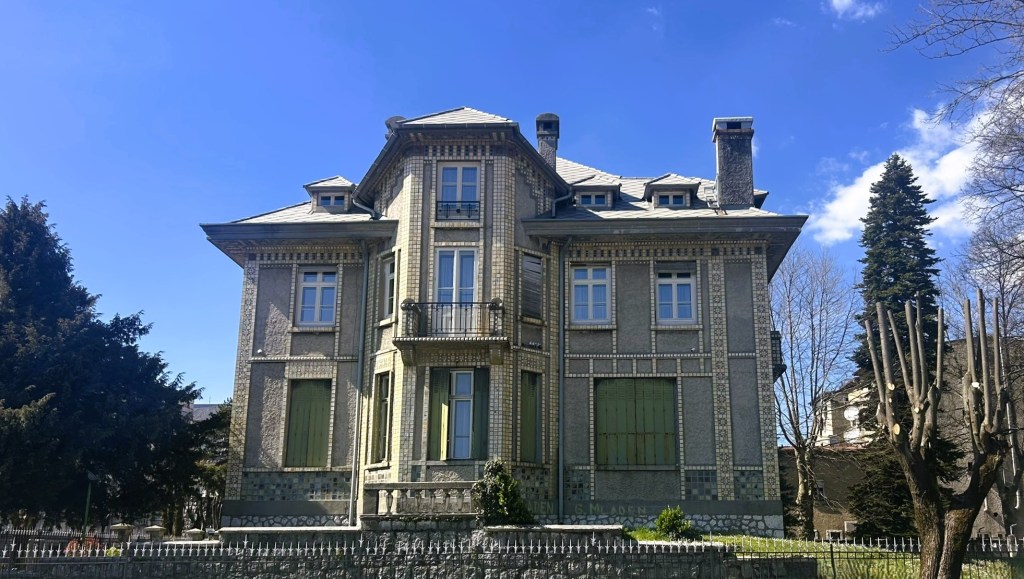 A historic three-storey house with intricate stonework, large windows, and green shutters, set against a blue sky.