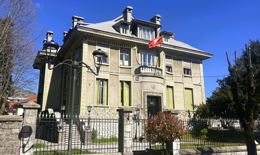 A historic two-storey building with a tiled roof, featuring large windows and a balcony. The exterior is adorned with decorative tiles and is surrounded by a wrought iron fence. A red flag is displayed prominently at the front.