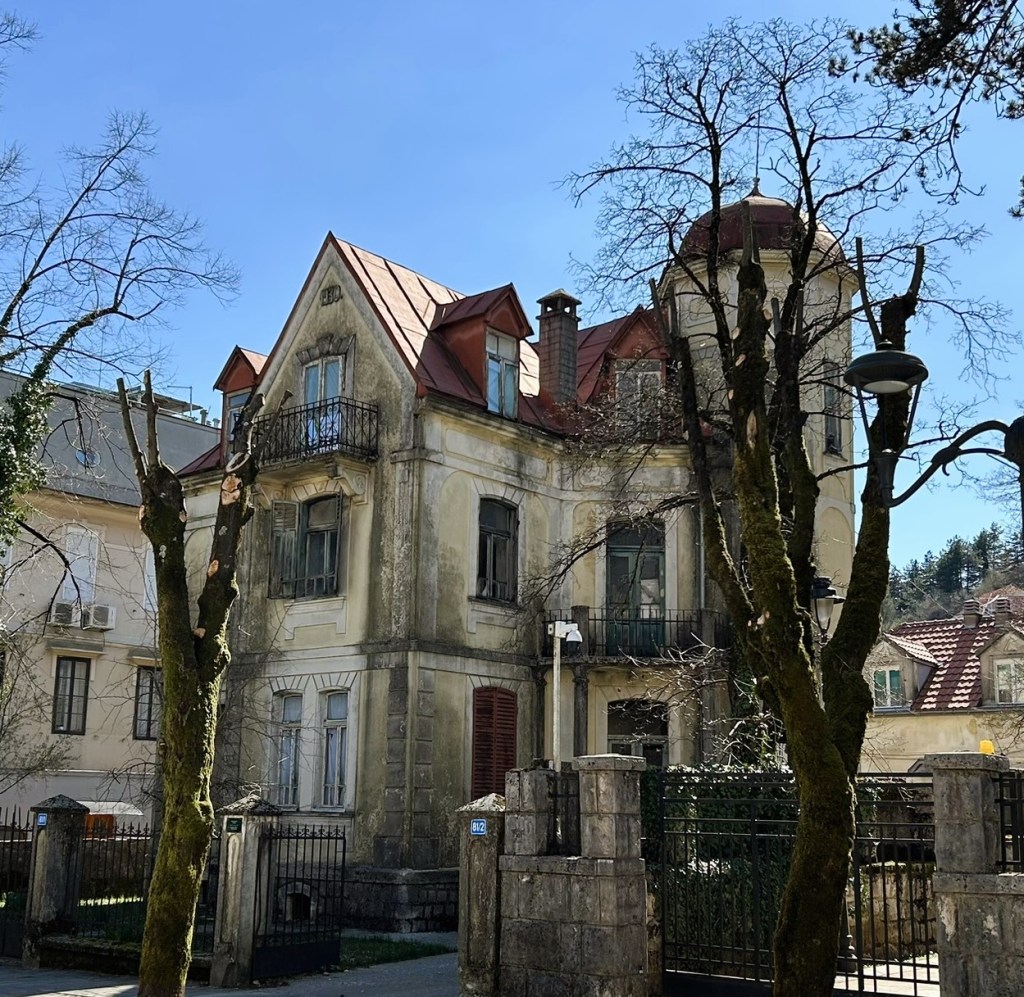 An old, dilapidated house with a red metal roof and a turret, surrounded by bare trees and a wrought iron gate.