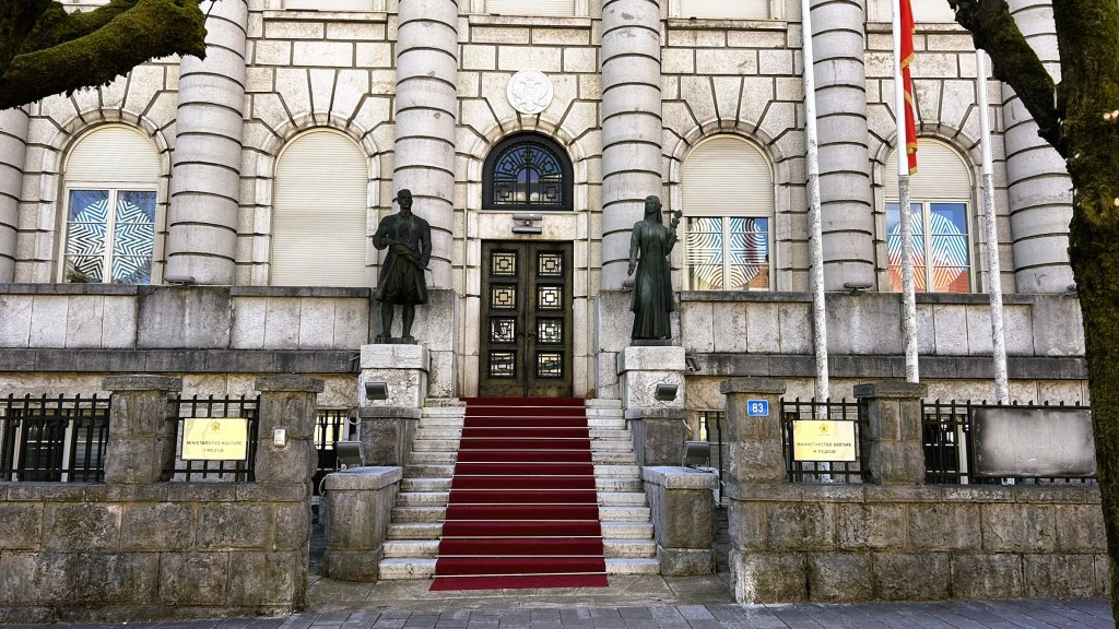 Front entrance of a historic building featuring two statues, a staircase with red carpet, and decorative stone architecture.