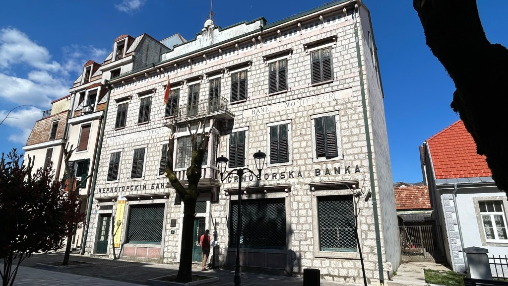 Facade of a historic bank building with stone architecture, displaying the name 'Crnogorska Banka' and decorative elements, set against a clear blue sky.