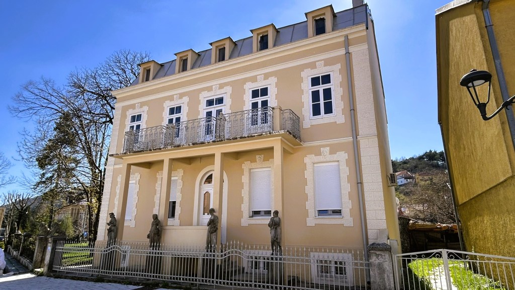 A light-coloured, three-storey building with a balcony, featuring decorative stonework and statues at the front, set against a clear blue sky.