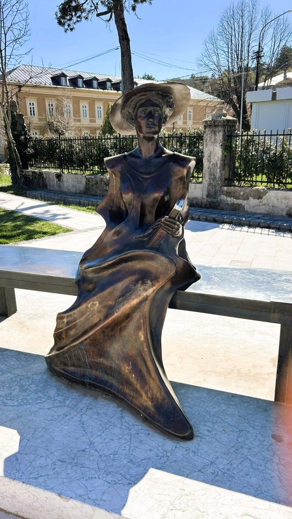 A bronze statue of a woman sitting on a bench, wearing a wide-brimmed hat and holding a book, set in a park-like environment with trees and buildings in the background.