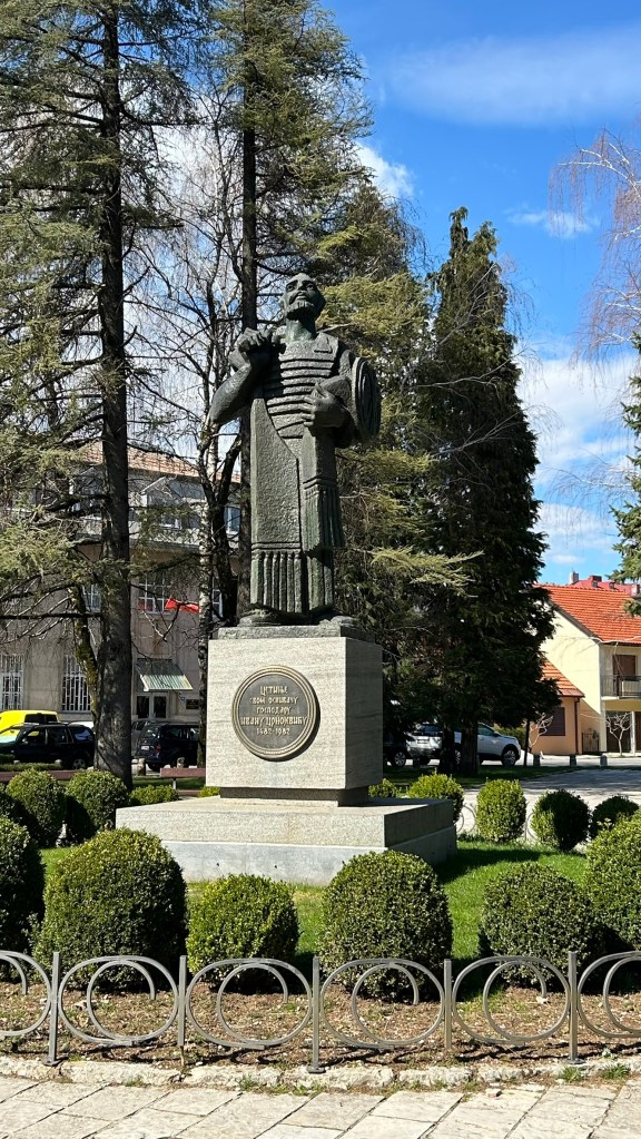 A statue of a historical figure in a green robe, holding a lyre, situated in a park surrounded by trimmed bushes and trees, with a blue sky in the background.