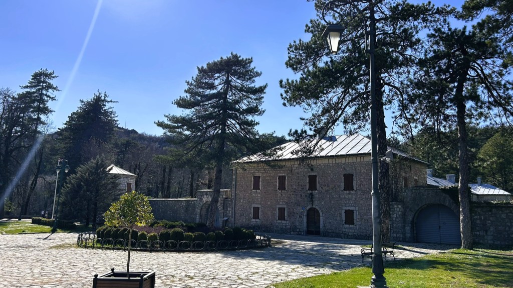 A stone building surrounded by trees and greenery, featuring a paved pathway and lampposts in a sunny outdoor setting.