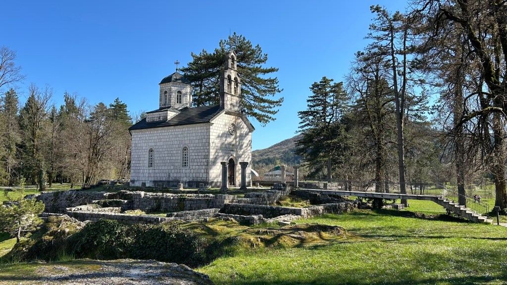 A stone church surrounded by trees and greenery on a clear blue sky day, with historical ruins in the foreground.
