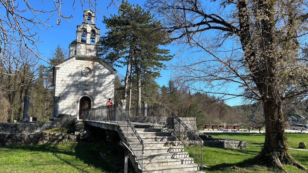 A small stone church with a bell tower, surrounded by trees and greenery, featuring a stairway leading to its entrance.