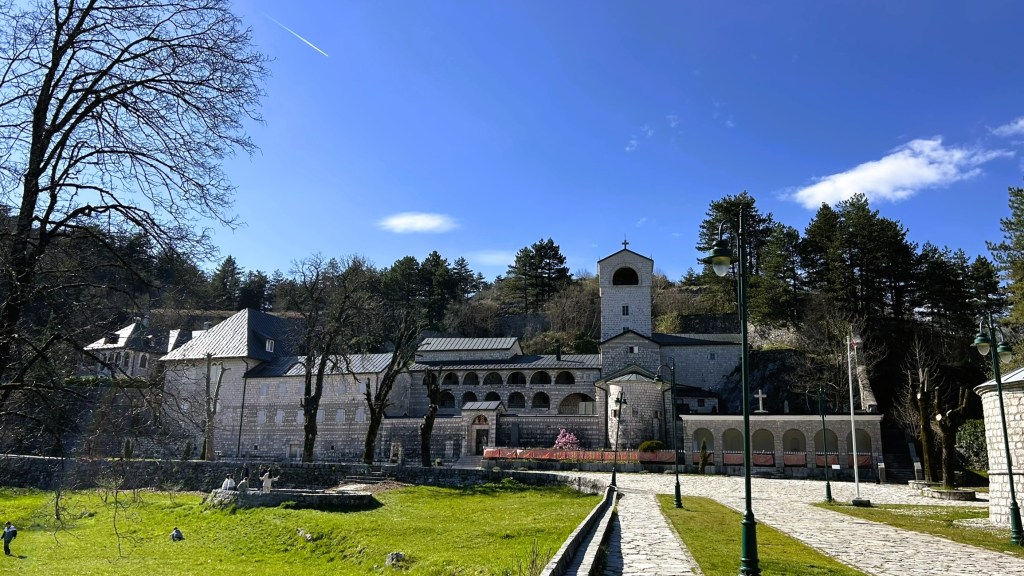 A scenic view of a stone monastery surrounded by greenery under a clear blue sky, featuring a bell tower and multiple buildings with contrasting roofs.
