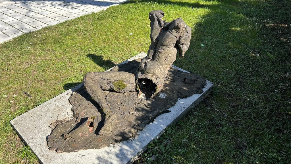 Sculpture of a humanoid form partially emerging from a textured surface, placed on a stone slab in a grassy area.
