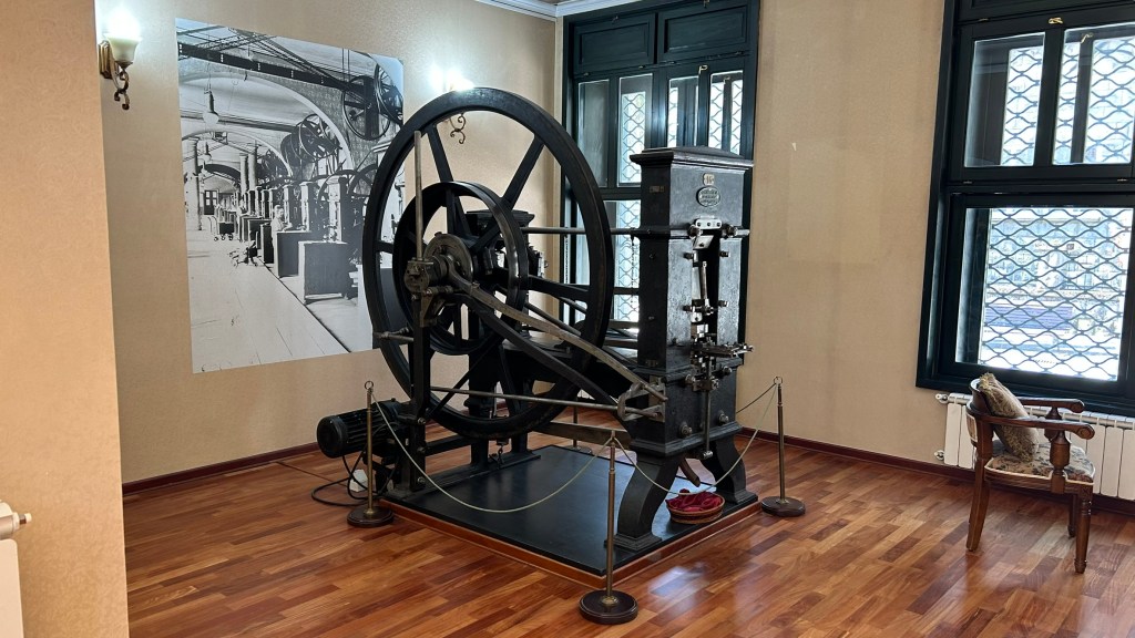 An antique printing press displayed in a museum setting, featuring a large wheel and machinery, with a black and white photograph on the wall depicting an old factory environment.