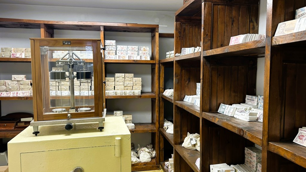 A vintage interior featuring wooden shelves filled with stacks of banknotes, alongside a glass-fronted weighing scale and a secure storage box.