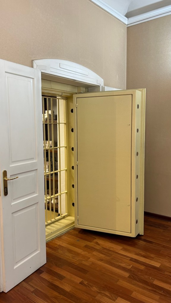 A large, open safe with a yellow door positioned in a room with wooden flooring. A white door is seen on the left, leading to the safe compartment.