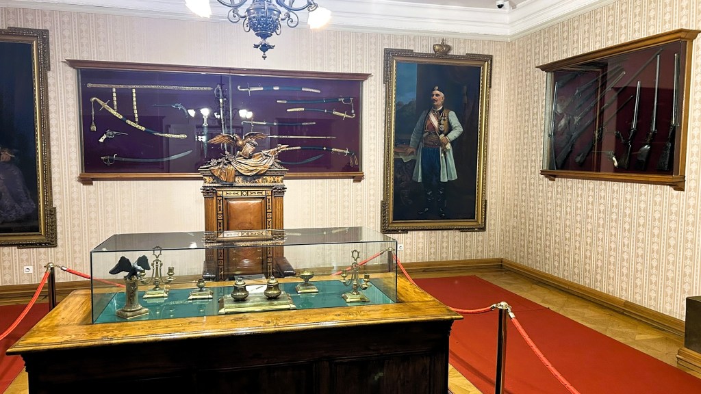Interior of a museum room displaying historical weapons and artifacts, including swords and firearms, with a glass display case at the centre and framed portraits on the walls.