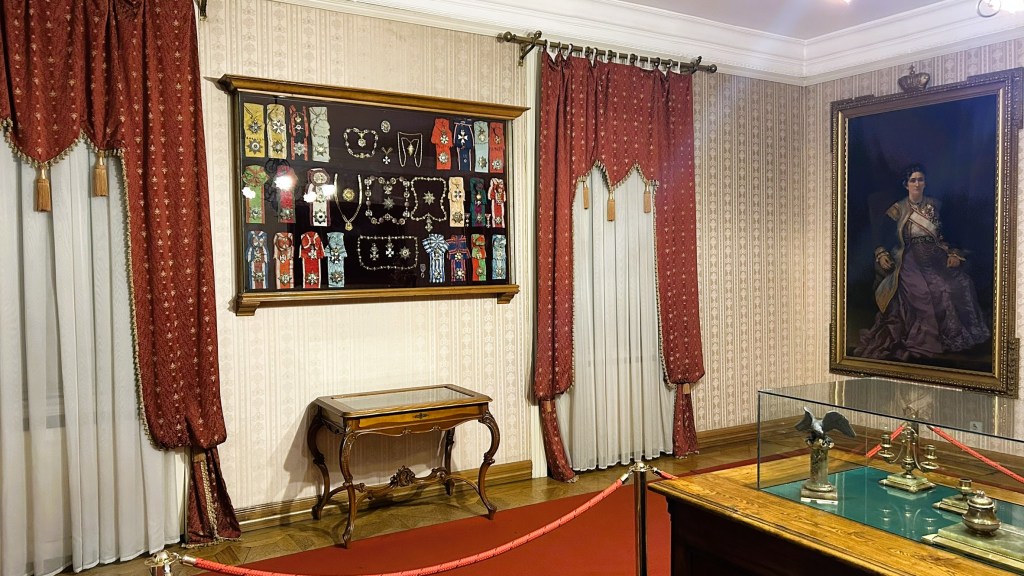 Interior of a museum room featuring decorative curtains, a display of jewellery on the wall, a wooden table, and a glass case with artifacts, along with a large painting of a woman in an elegant gown.