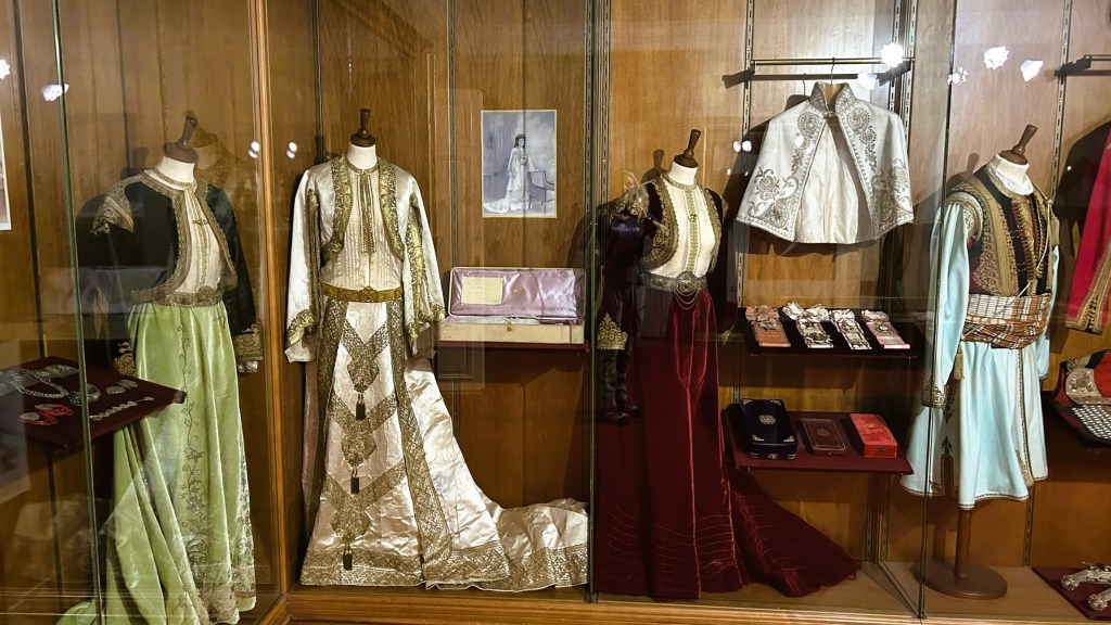 Display of traditional garments in a glass case, featuring ornate dresses and accessories, showcasing cultural attire.