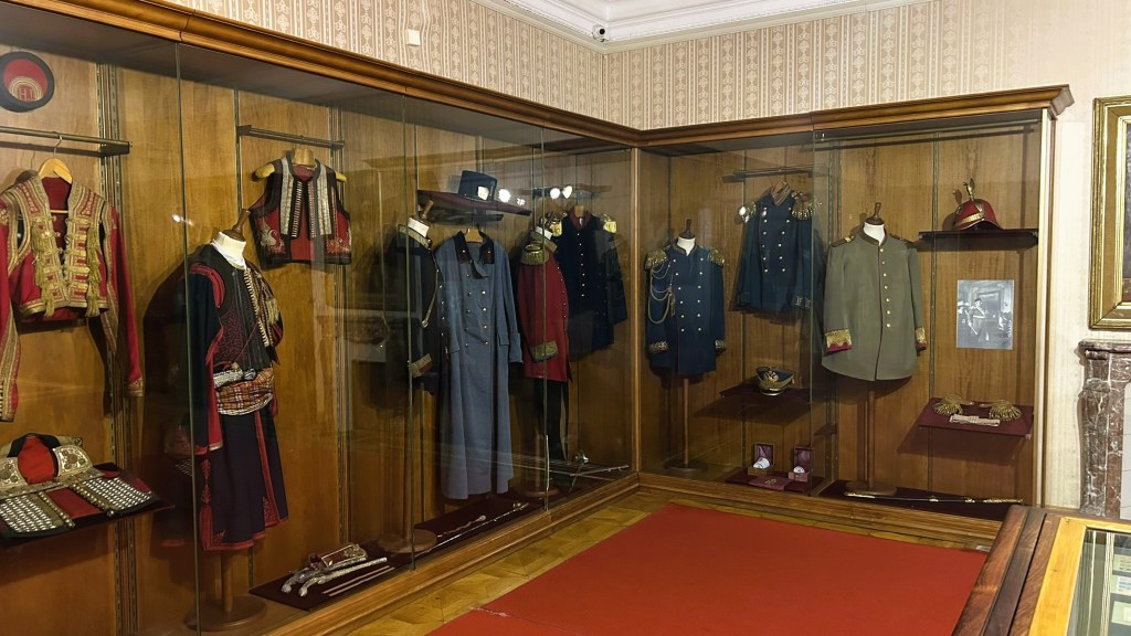 A display case showcasing various historical military uniforms and hats, featuring intricate designs and decorations, set against a wooden background.