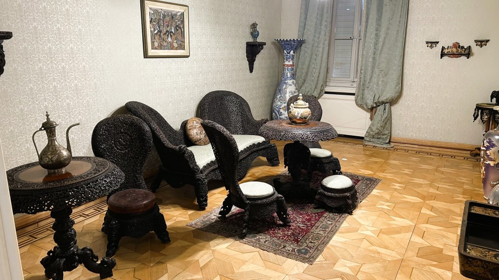 A traditional room featuring ornate wooden furniture, including two chairs, a table, and decorative stools, with intricate carvings. The floor is made of parquet, and there is a patterned rug. The walls are adorned with light-coloured wallpaper and a framed painting, while heavy curtains drape beside a window.