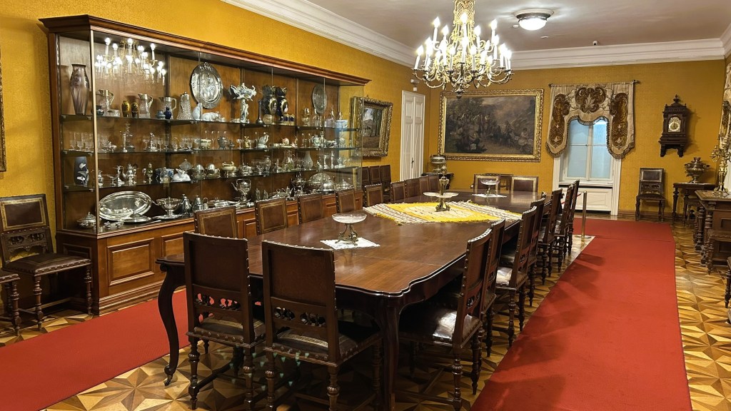 A lavish dining room featuring a large wooden table surrounded by chairs, a glass display cabinet filled with silverware, an ornate chandelier, and rich yellow walls.