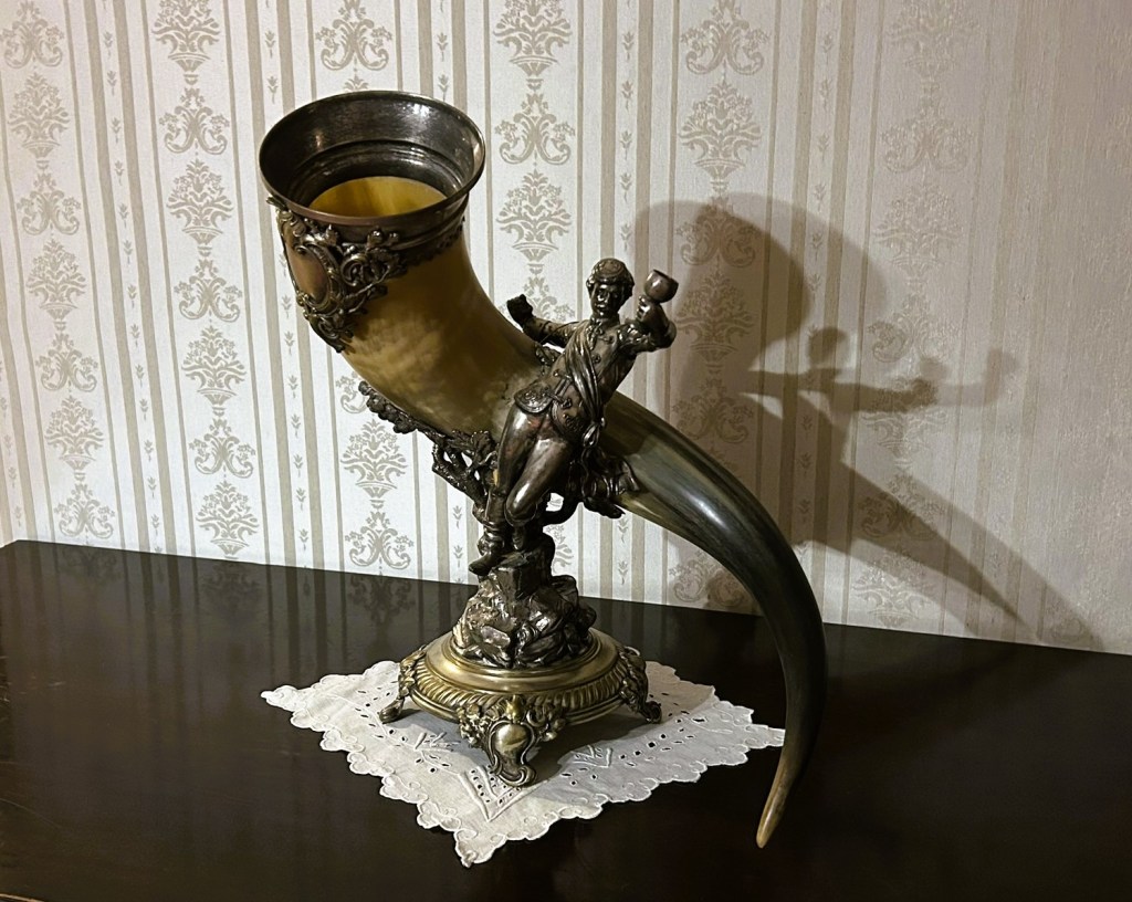 A decorative horn featuring a detailed figure of a soldier, resting on an ornate base. The background includes a patterned wallpaper.