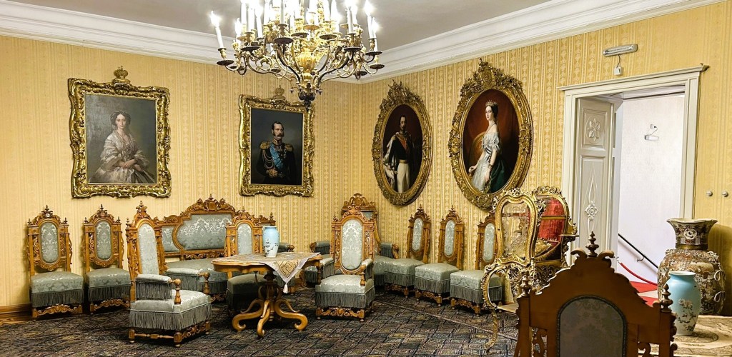 An elegantly furnished room featuring ornate golden-framed portraits on the walls, vintage upholstered chairs arranged around a decorative table, and an elaborate chandelier overhead.