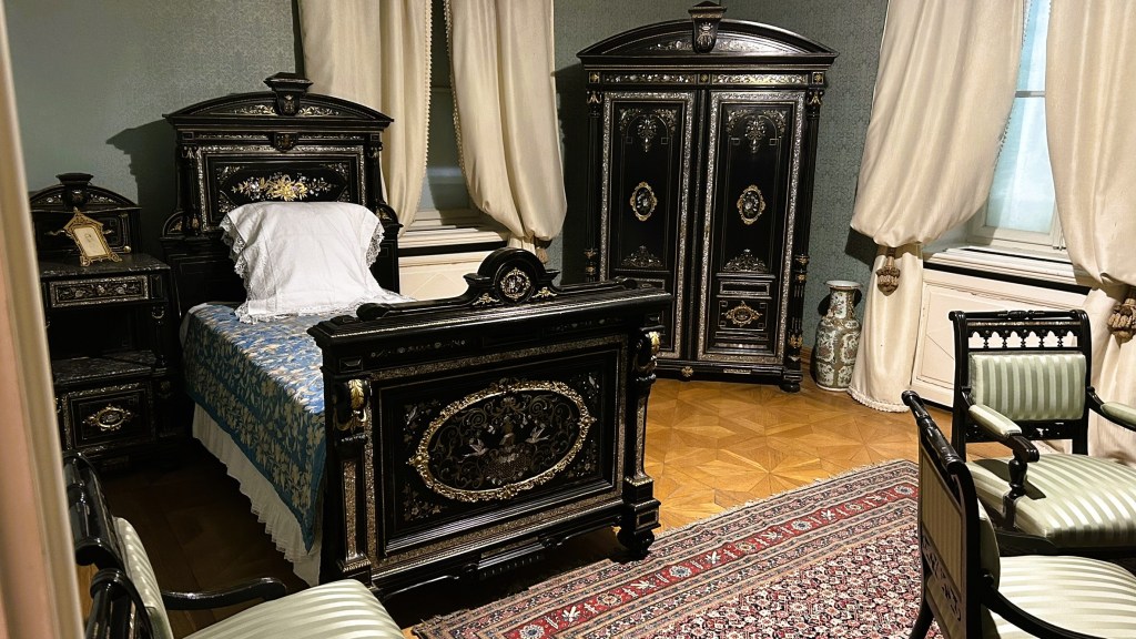 A vintage bedroom featuring an ornate black wooden bed with a blue and white patterned coverlet, a matching bedside table, a tall wardrobe, and two elegant chairs. There are sheer curtains covering the window, and a decorative rug on the floor.