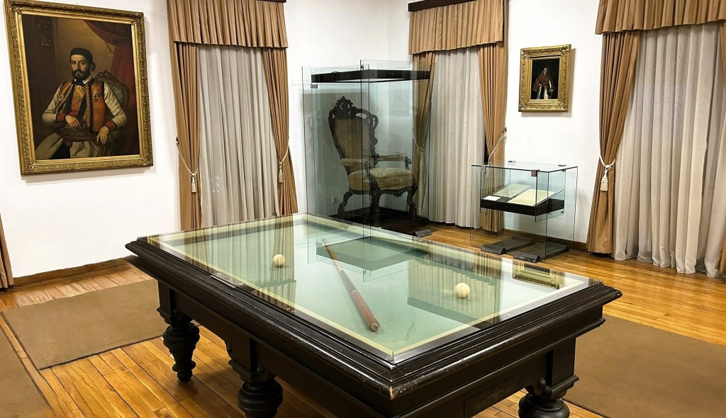 An interior view of a historical room featuring a large wooden table with a glass top, two balls, and a wooden rod. Behind the table is a decorative chair and two paintings on the walls, one of a seated man in historical attire and the other of a similarly dressed figure. The room has cream-coloured curtains and wooden flooring.