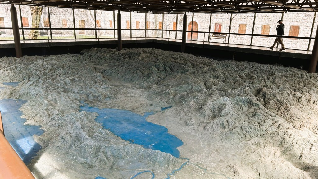 A large topographical relief model depicting a mountainous landscape with a blue lake, viewed from above with a person walking in the background.