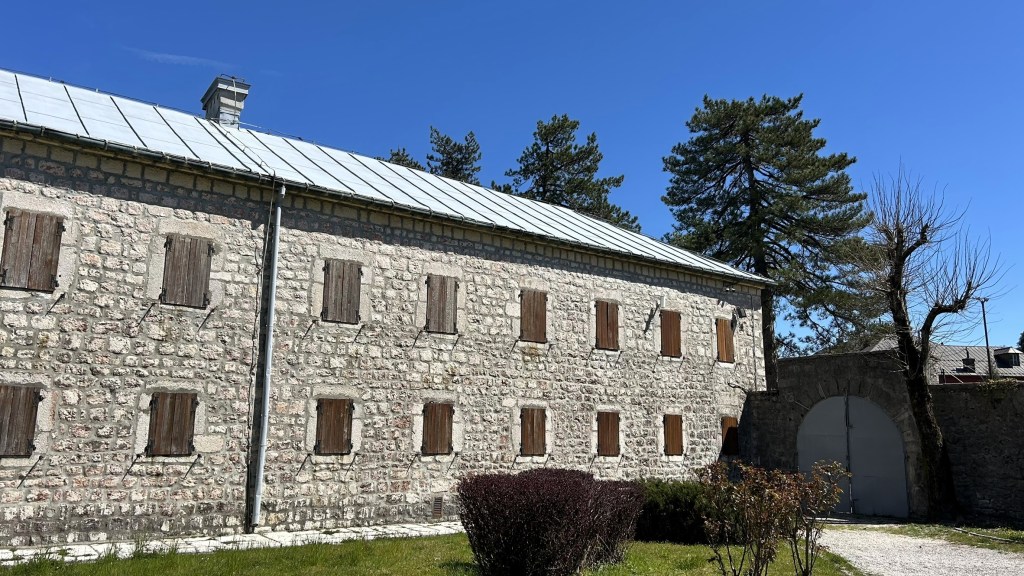 A stone building with wooden shutters and a grey metal roof, surrounded by grass and trees under a clear blue sky.